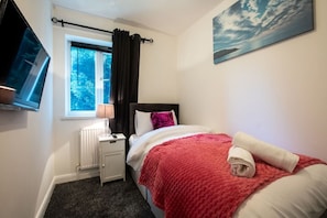 A bedroom with a bed, a window with curtains, a radiator, a bedside table with a lamp, and a painting on the wall.