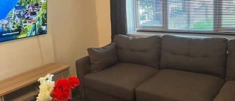 A living room with a dark brown sofa, a wooden coffee table with a vase of flowers, and a large window with blinds.