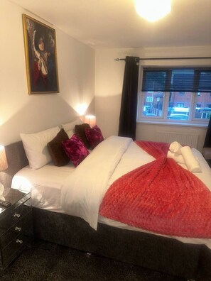 Hotelkamer met een bed, kussens, dekbed, schilderij, lamp, raam met gordijnen en uitzicht op gebouwen.