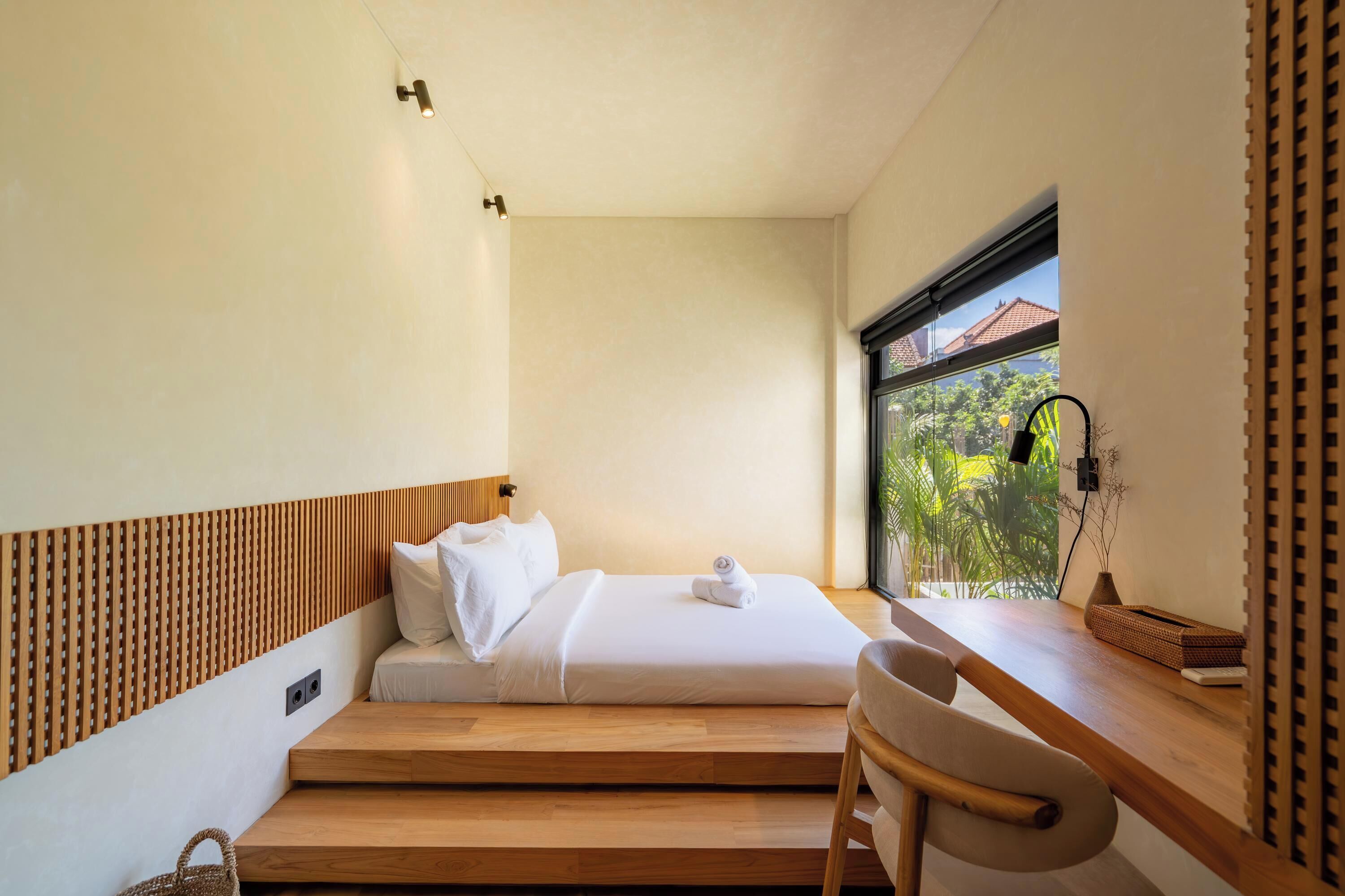 A modern bedroom with a bed, wooden desk, and a window with a view of greenery.