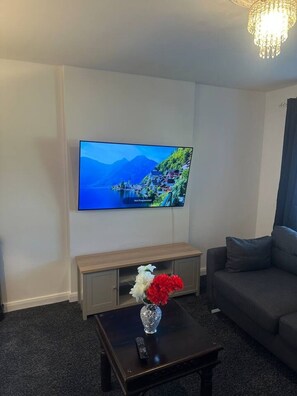 A living room with a flat-screen TV mounted on the wall, a wooden cabinet, a coffee table with a vase of flowers, and a gray sofa.