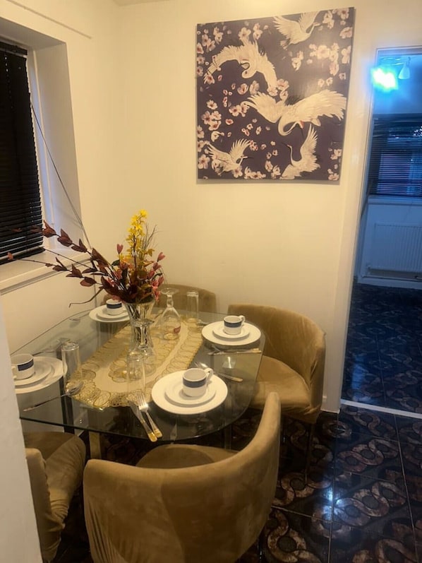 A dining area with a glass table, beige chairs, a vase with flowers, and a wall-mounted artwork.