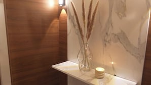 A bathroom with a marble wall, wooden paneling, a white shelf with a vase and candle, and a light fixture. - Cozzy Inn (Oran)