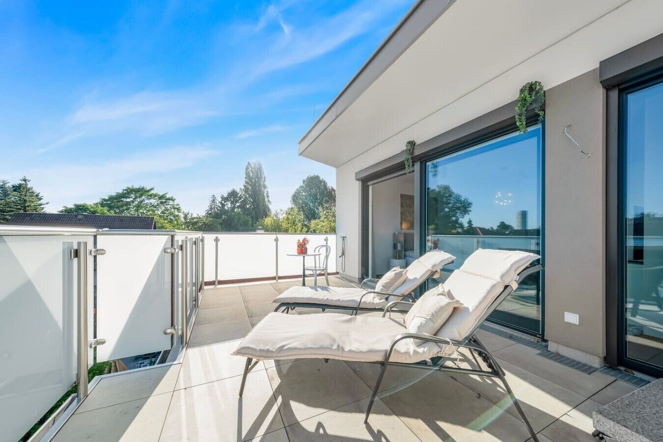 A rooftop terrace with three sun loungers, a glass door, and a view of trees and buildings.