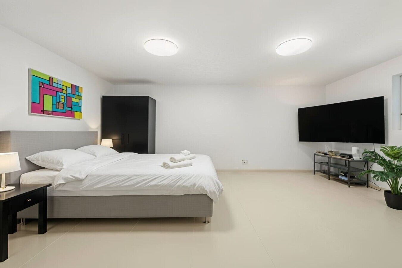 Een moderne hotelkamer met een bed, nachtkastje, lamp, kunstwerk en een televisie aan de muur.