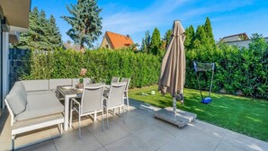 Eine Terrasse im Freien mit Esstisch und Stühlen, einem schattenspendenden Sonnenschirm und einem gepflegten Rasen.