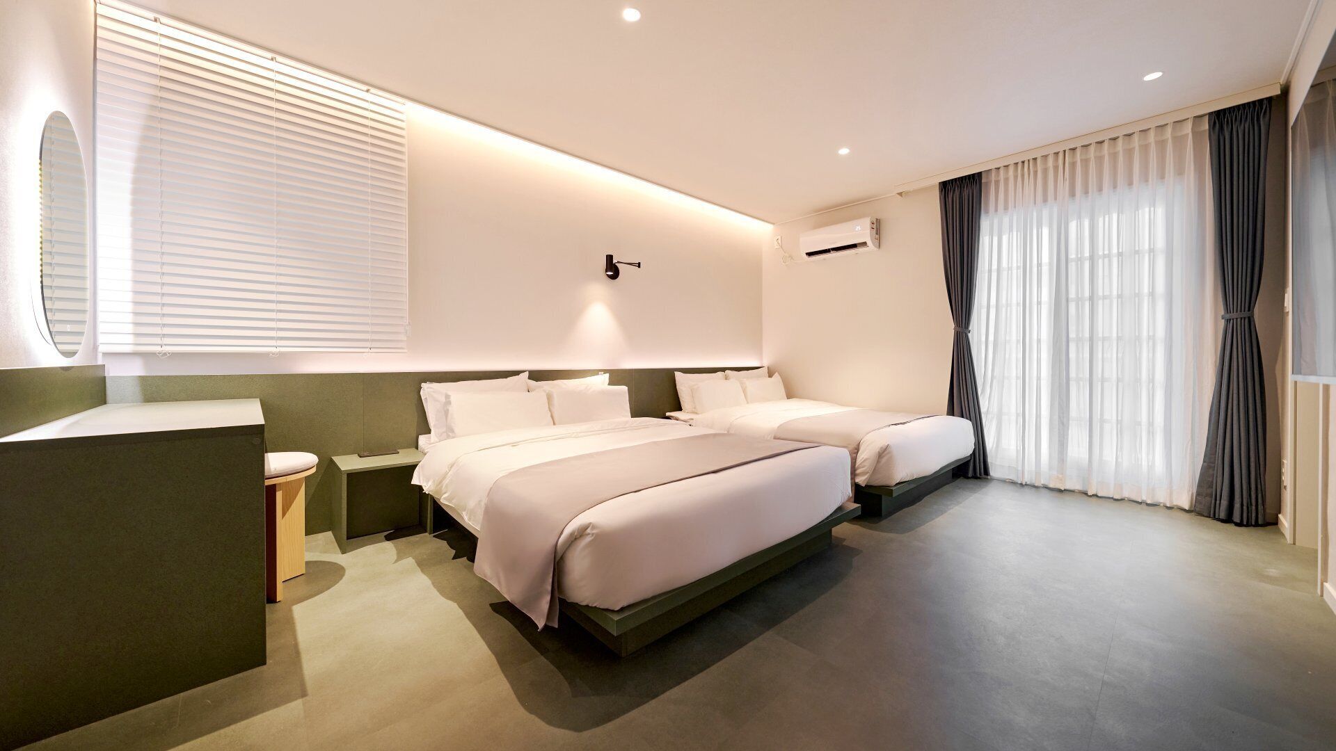 A hotel room with two beds, a bedside table, a mirror, an air conditioning unit, and a window with curtains.