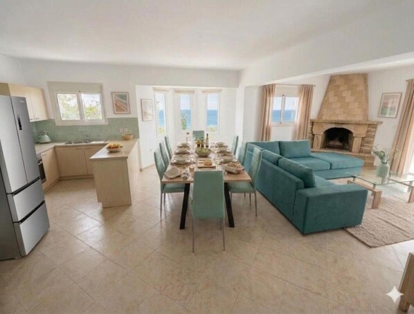 A spacious living area with a dining table set for four, a teal sofa, a fireplace, and a kitchen with a refrigerator.
