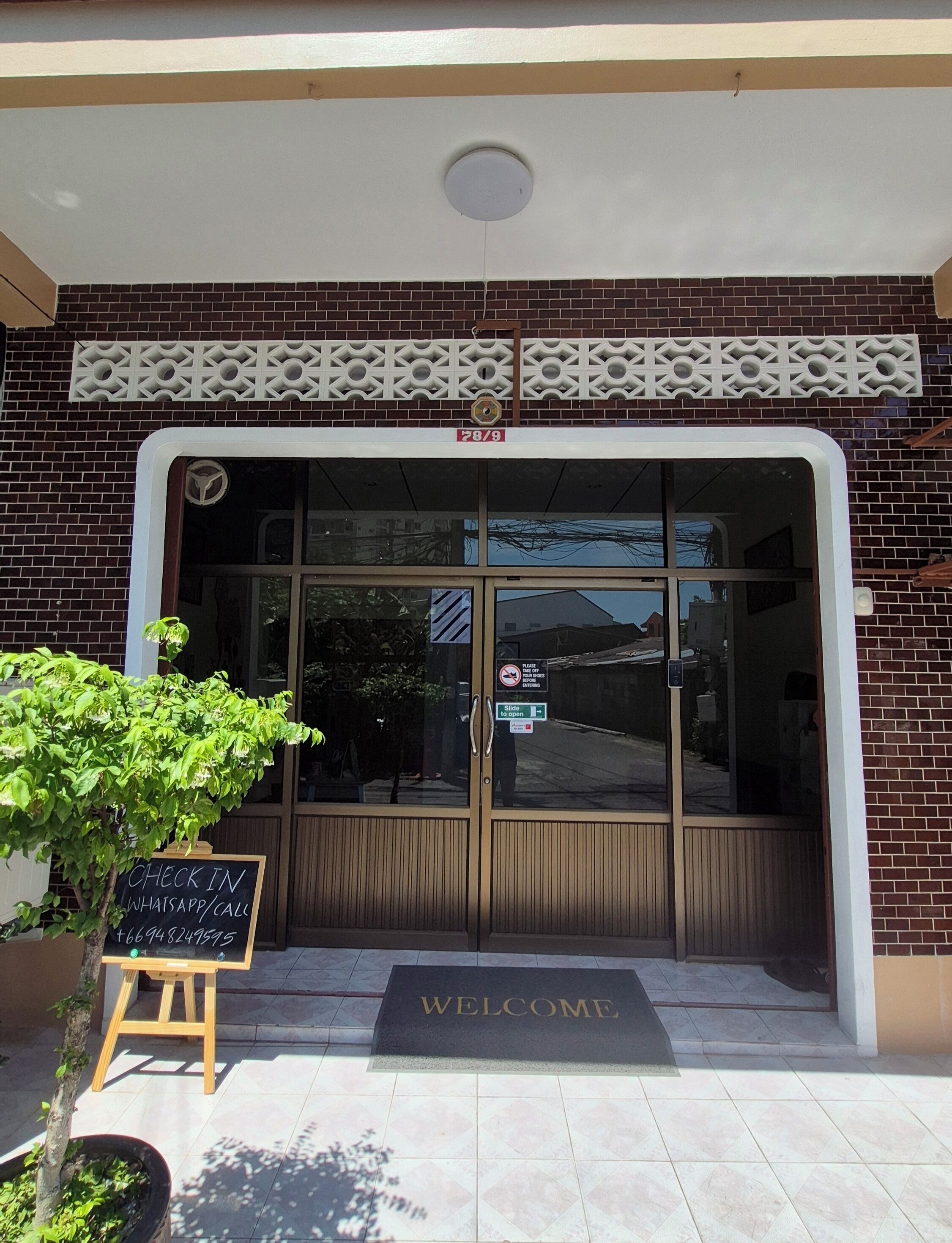 A building entrance with a glass door, a 'WELCOME' mat, and a sign offering check-in via WhatsApp or call.