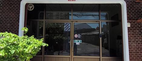 A building entrance with a glass door, a 'WELCOME' mat, and a sign offering check-in via WhatsApp or call.