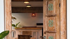 Uma porta de madeira aberta com padrões verdes detalhados, dando acesso a um quarto com mesa e cadeiras de madeira, e um candeeiro pendente.