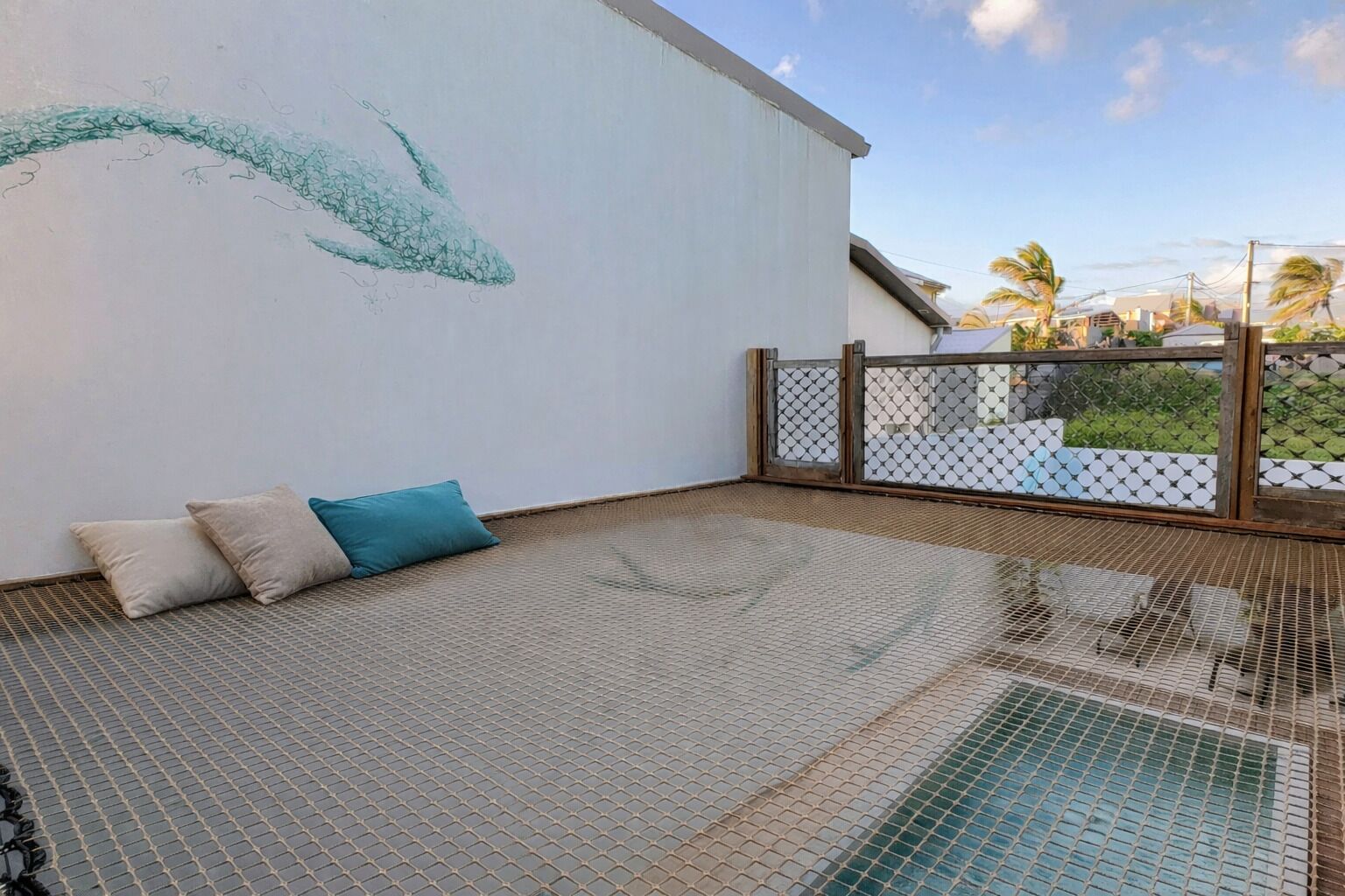 A balcony with a mesh floor, a white wall with a green abstract design, and a view of palm trees and buildings.