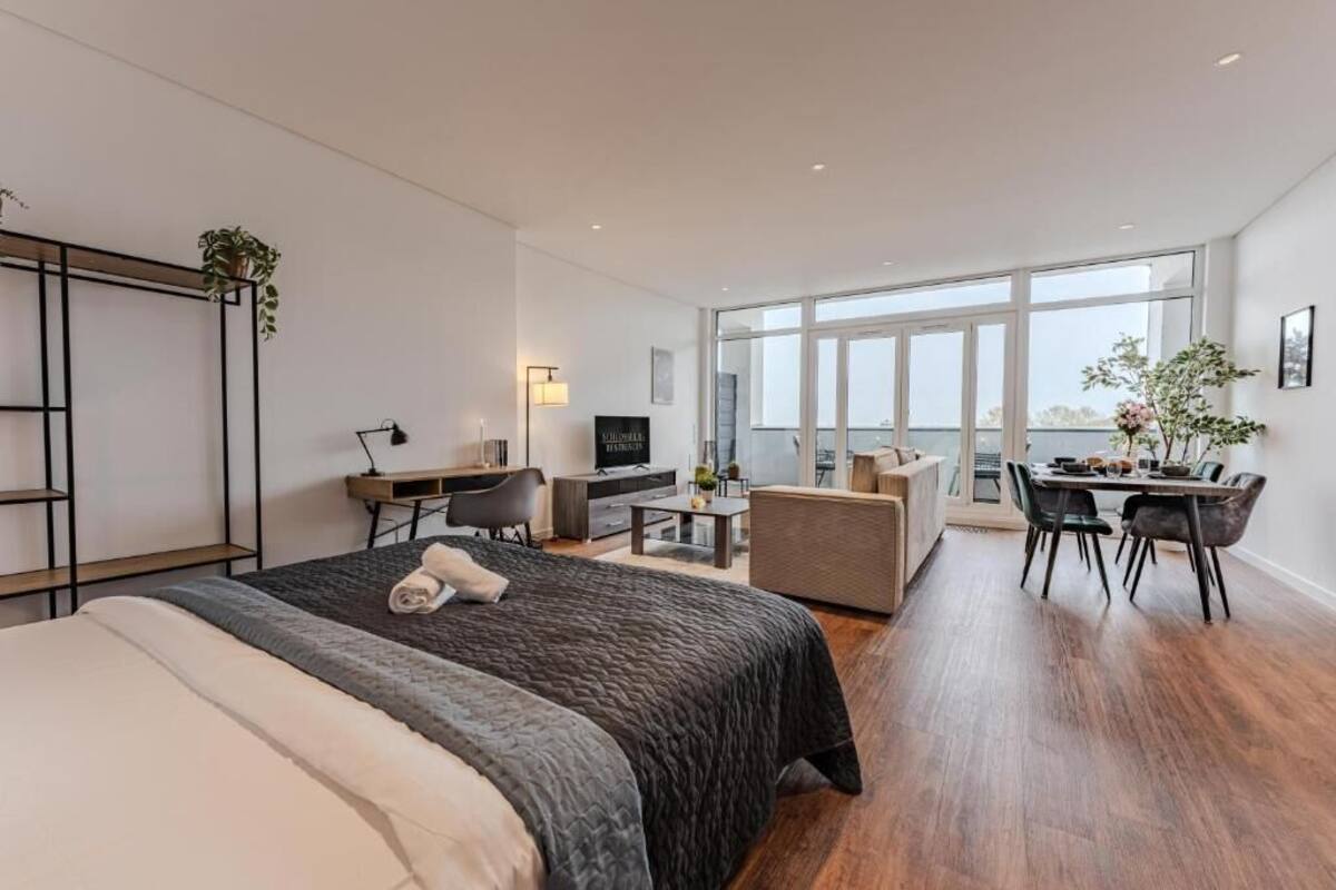 A modern hotel room with a large bed, a desk, a dining area, and a balcony with a view.