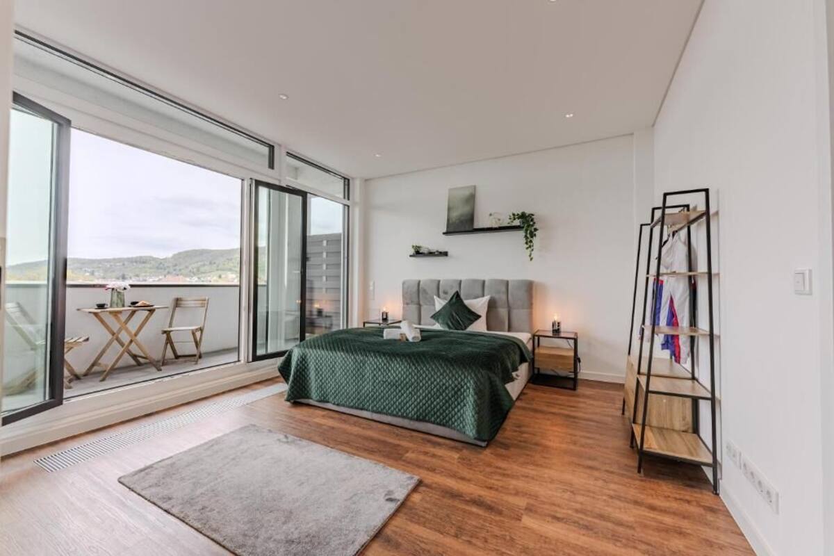 A bedroom with a large sliding glass door leading to a balcony, a bed with a green quilt, a bedside table, and a wooden shelf unit.