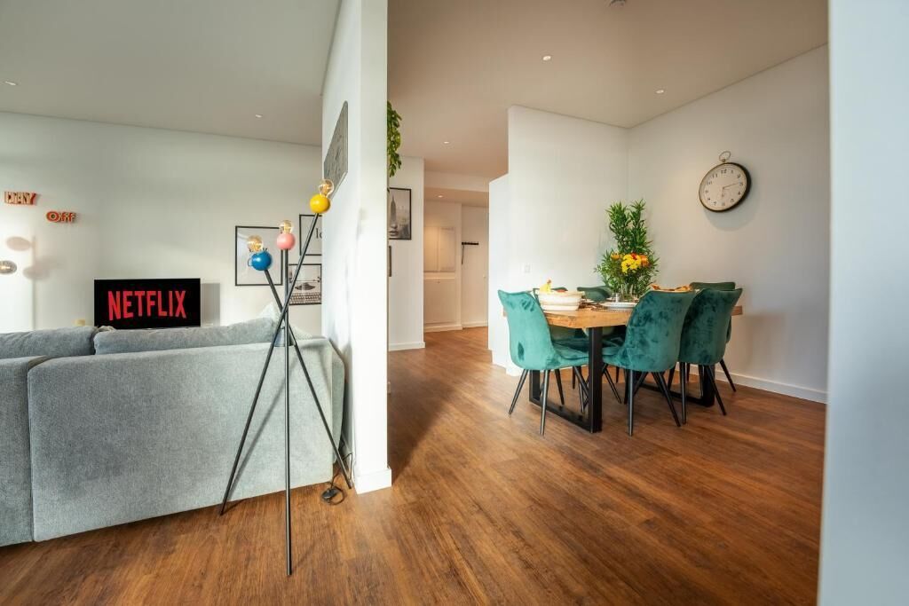 Una sala moderna con un sofá gris, una mesa de comedor con sillas color turquesa, un trípode y un cartel de 'NETFLIX'.