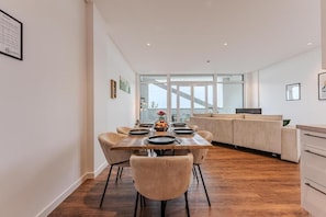A modern dining area with a long table, chairs, and a kitchen in the background.