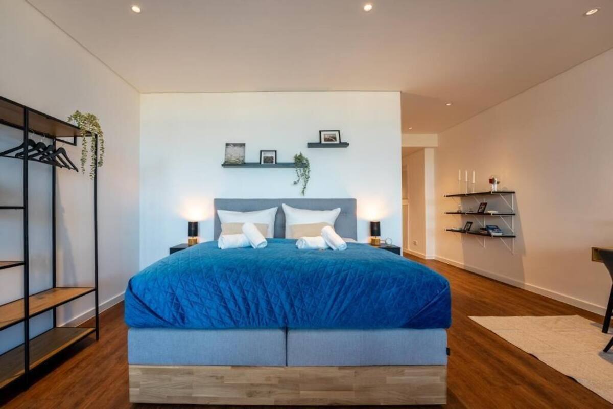 A modern bedroom with a large bed, bedside tables, a wall-mounted shelf with pictures, and a plant.