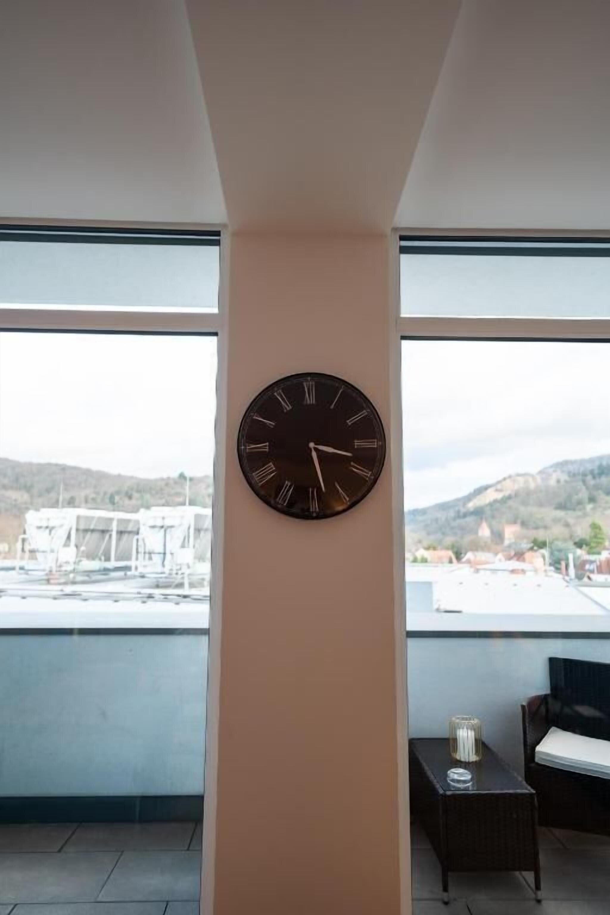 A room with a large wall clock, a small table with a candle, and a view of a cityscape through large windows.