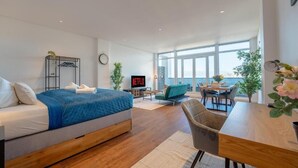 Una habitación de hotel moderna con una cama grande, zona de comedor, televisor y vista al mar.