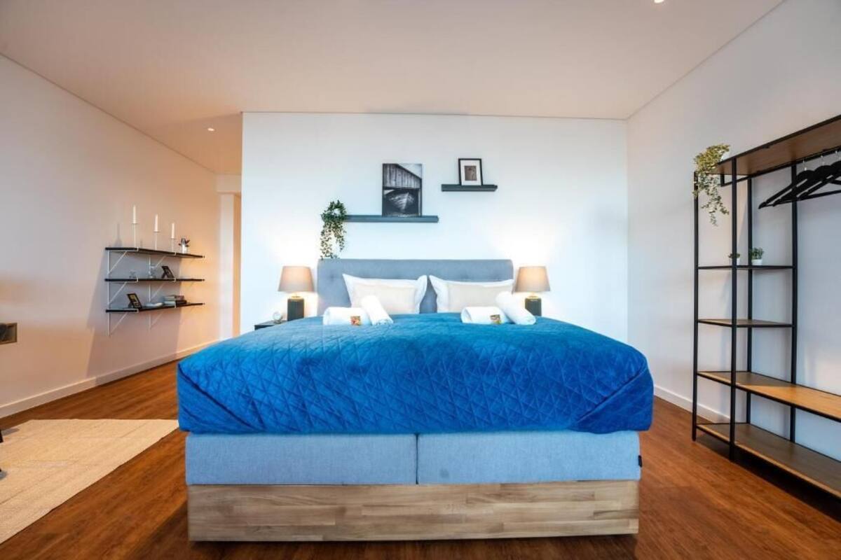 A modern bedroom with a large bed, bedside lamps, a wall-mounted shelf with decorative items, and a built-in wardrobe.