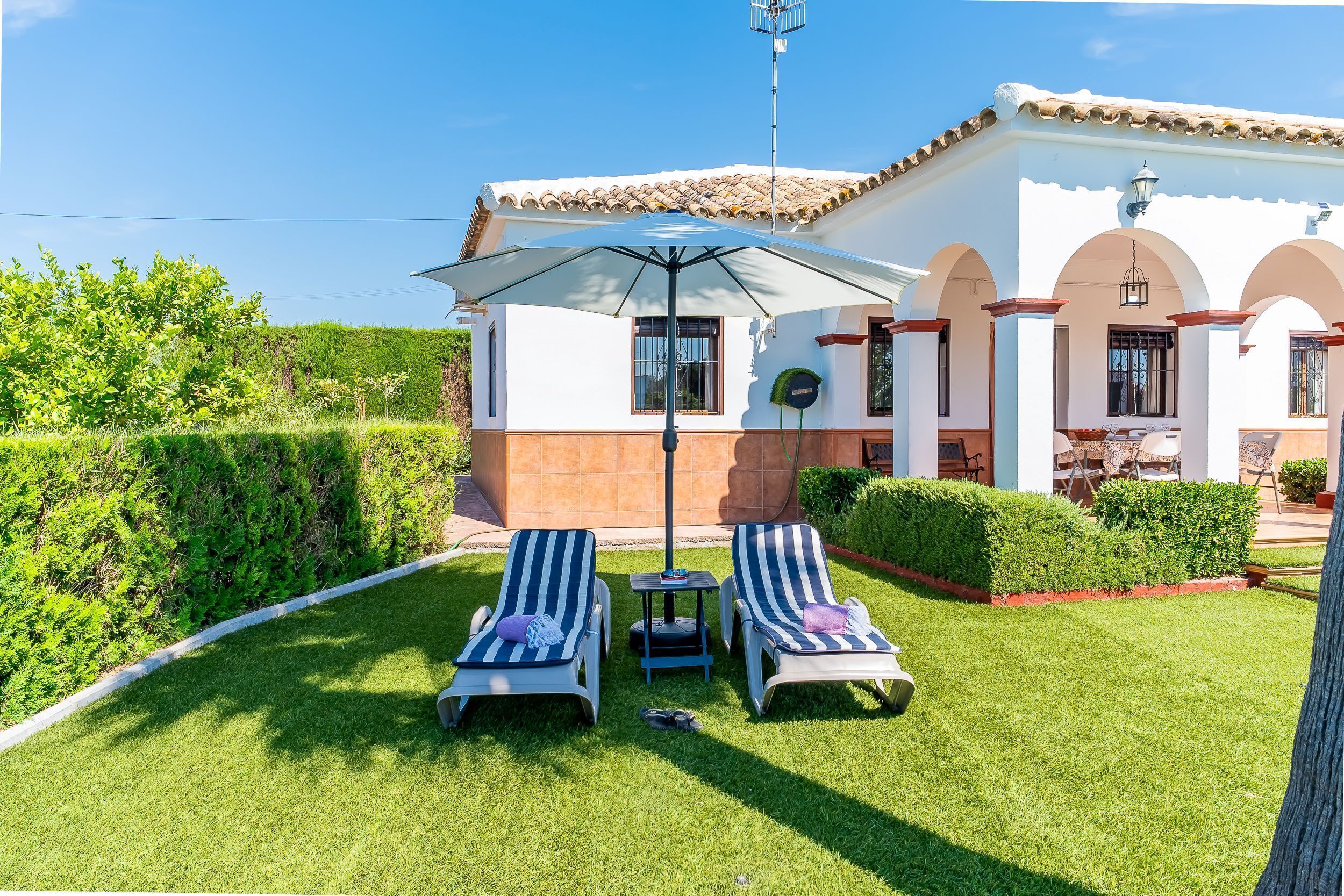 A house with a tiled roof, a white exterior, and a well-maintained lawn with two lounge chairs and an umbrella.