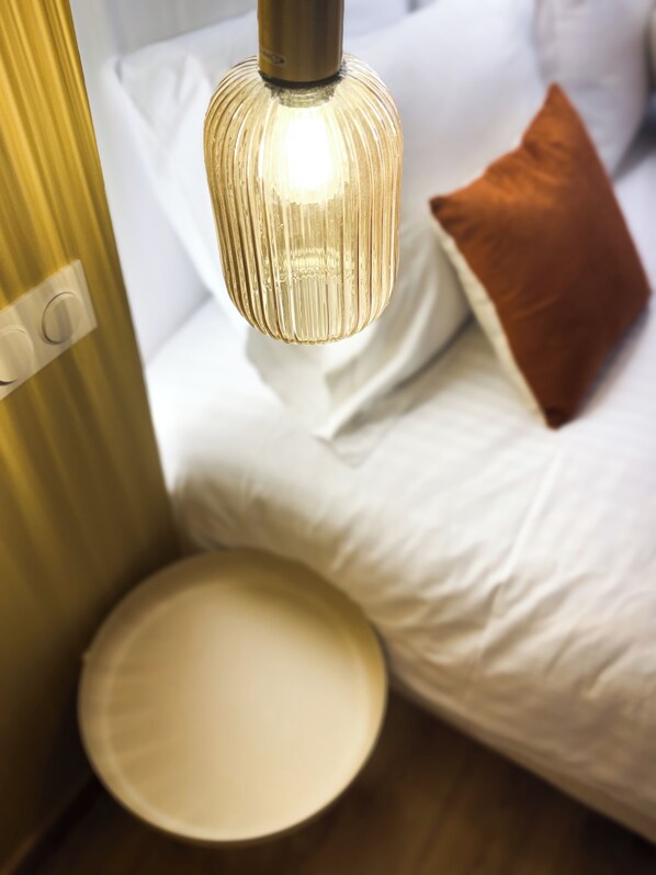 Un lit avec une literie blanche et un oreiller marron, une lampe de chevet et une table ronde.