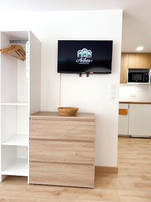 Una camera moderna con TV a parete, un mobiletto contenitore in legno e un angolo cottura con microonde.