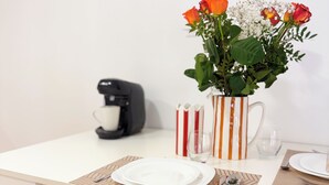 Ein Essbereich mit einem weißen Tisch, einer Kaffeemaschine, einer gestreiften Tasse, einer Vase mit Blumen und einem roten Stuhl.