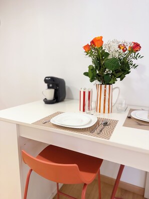 Zona pranzo con tavolo bianco, macchina da caffè, tazza a righe, vaso con fiori e sedia rossa.