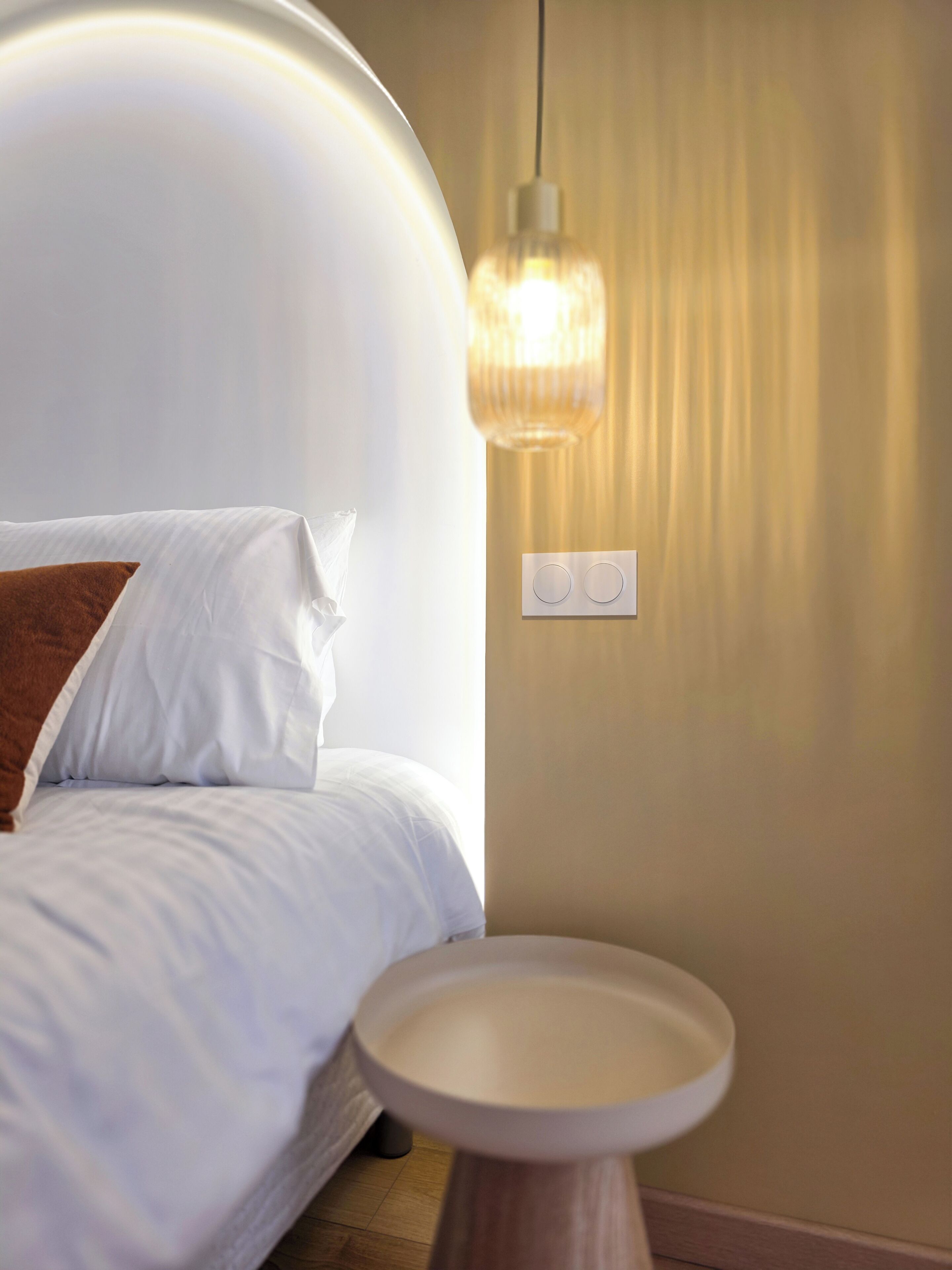 A neatly made bed with white linens and a brown pillow, a round wooden side table, and a wall-mounted light fixture.