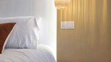 A neatly made bed with white linens and a brown pillow, a round wooden side table, and a wall-mounted light fixture.