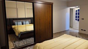 A bedroom with a large mirror, a bed with a yellow duvet, a wooden wardrobe, and a hallway leading to a door with a blue window. - Stylish 3-bedroom apartment with AC in Nasr city, Near to Cairo Airport (Nasr City)