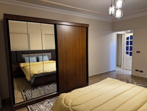 A bedroom with a large mirror, a bed with a yellow duvet, a wooden wardrobe, and a hallway leading to a door with a blue window. - Stylish 3-bedroom apartment with AC in Nasr city, Near to Cairo Airport (Nasr City)