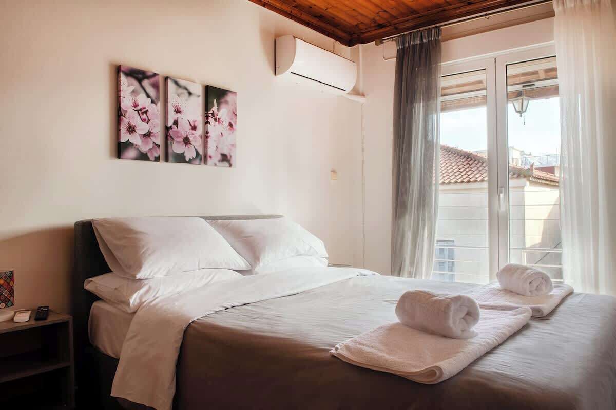 A hotel room with a bed, pillows, a nightstand, an air conditioning unit, and a window with curtains.