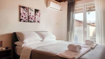 A hotel room with a bed, pillows, a nightstand, an air conditioning unit, and a window with curtains.