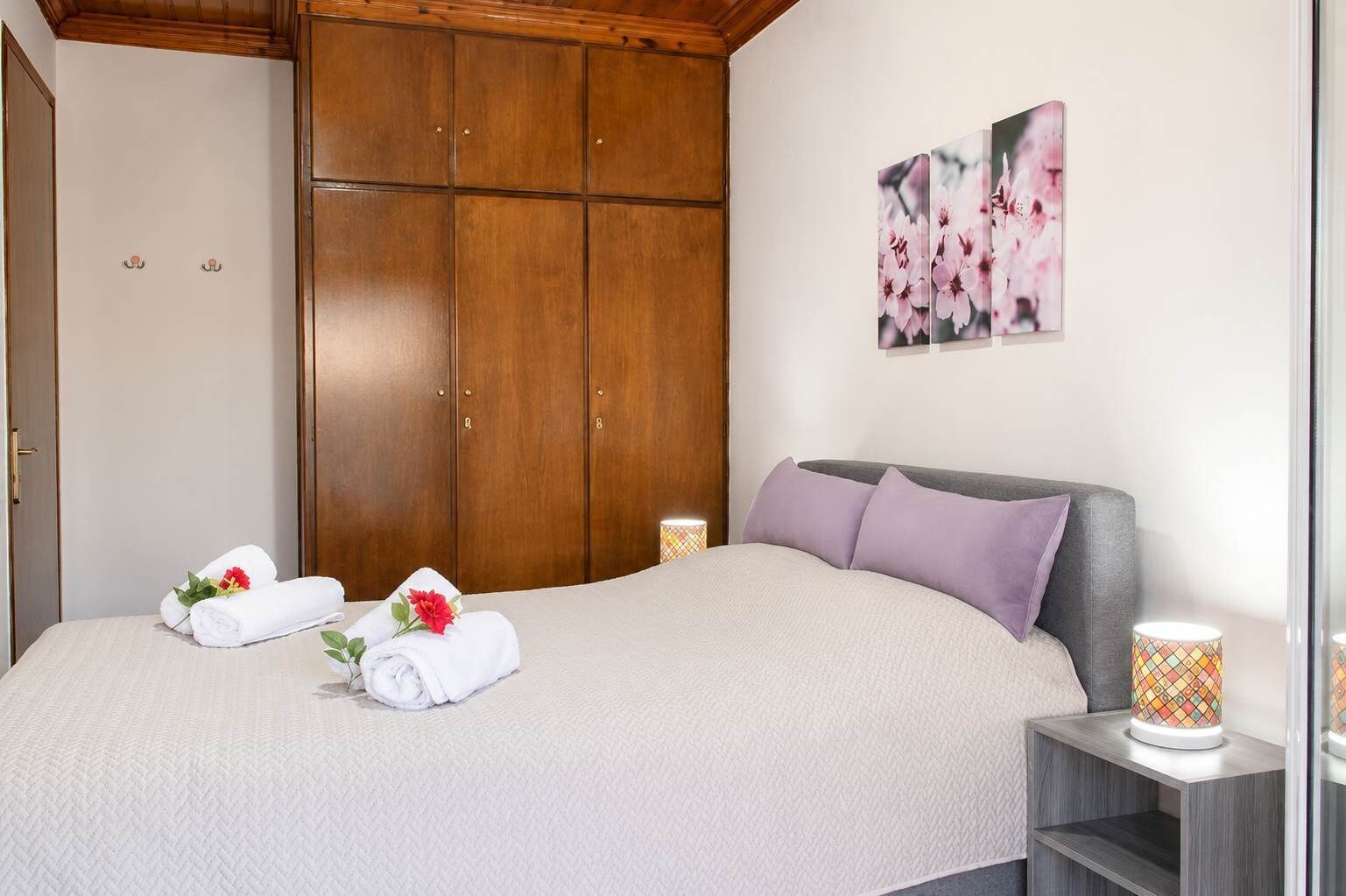 A hotel room with a bed, wooden wardrobe, and a wall-mounted picture of flowers.
