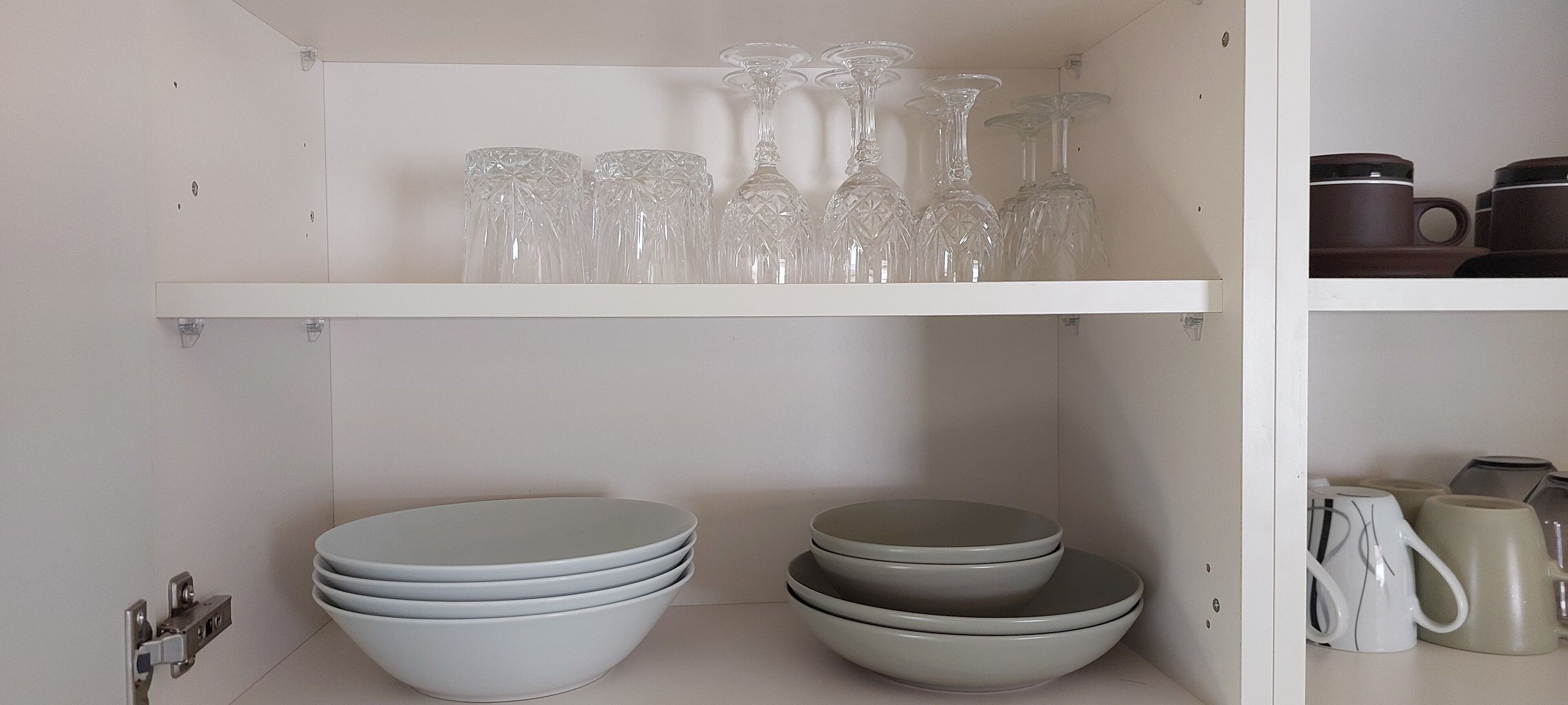 Ett kök med vita skåp, en vit hylla med genomskinliga glas och staplade vita skålar.