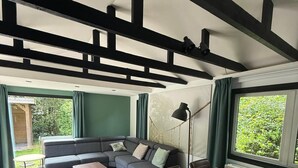 A modern living room with a sectional sofa, a wooden dining table, and large windows with curtains. - Intimate lodge (48 m²) for 2 people with full kitchen and spacious garden. Your four-legged friend is welcome too for an unforgettable Veluwe holiday. (Harderwijk)