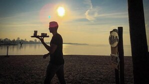 Une personne tenant un plateau avec des tasses et un panneau indicateur, dans un cadre côtier.