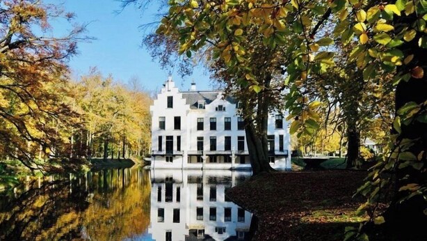 Un bâtiment à la façade symétrique, qui se reflète dans l’eau, et des arbres aux feuilles d’automne.
