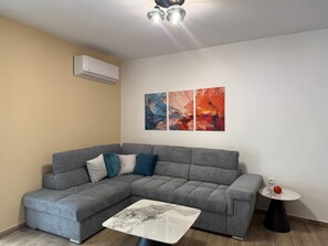 Ein Wohnzimmer mit einem grauen Ecksofa, einem kleinen runden Tisch mit einem roten Gegenstand und abstrakter Kunst an der Wand.