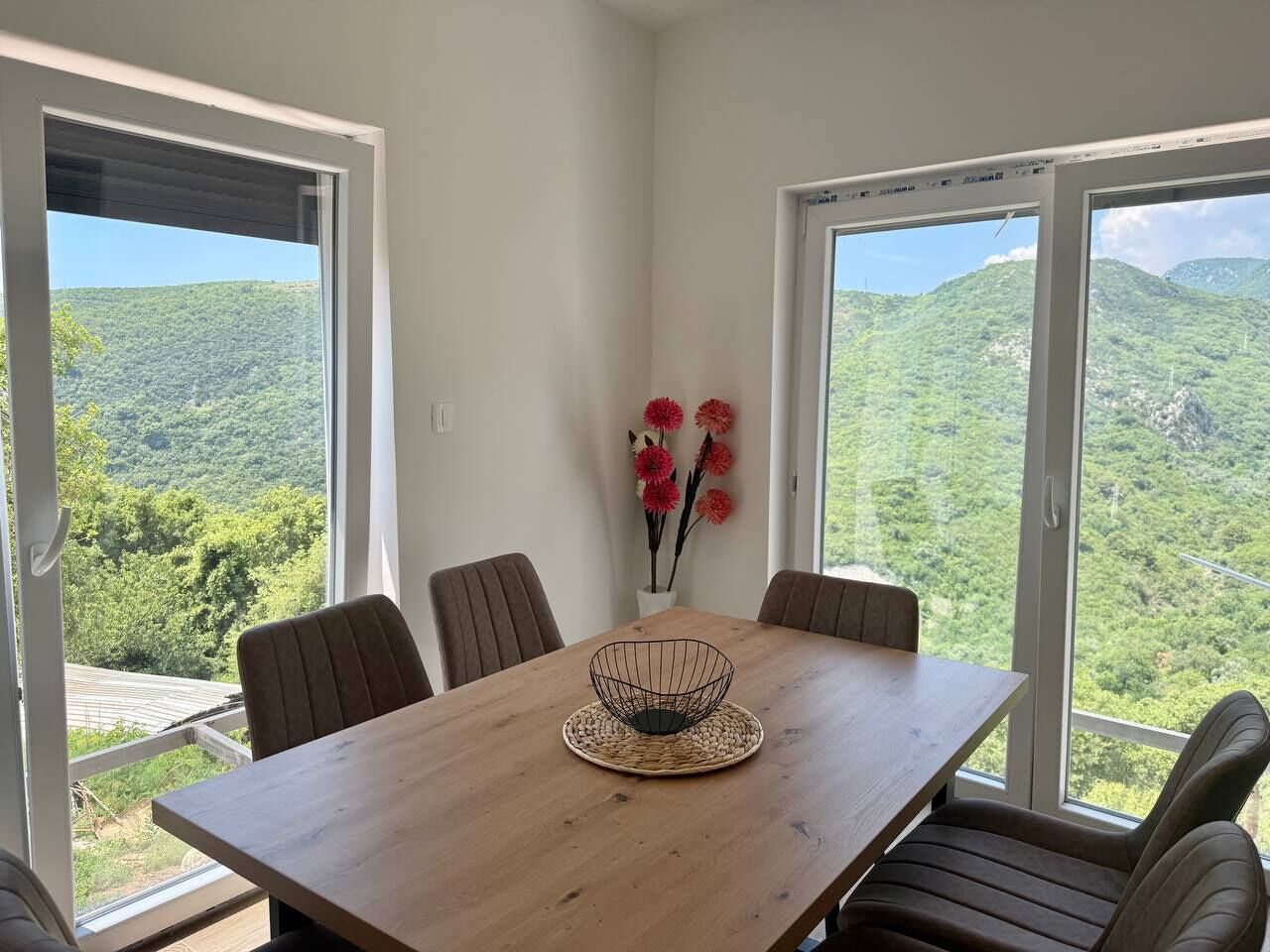 Un coin repas avec une table en bois, quatre chaises, un set de table en fibres tressées et un vase avec des fleurs rouges.