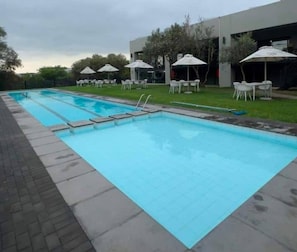 Ein Schwimmbecken mit mehreren Bahnen, umgeben von weißen Stühlen und Sonnenschirmen, mit einem modernen Gebäude im Hintergrund.