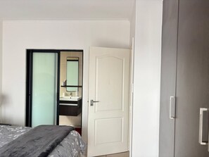 Ein Schlafzimmer mit einem Bett, einer weißen Tür, einem Spiegel und einem Badezimmer mit Waschbecken und Spiegel.