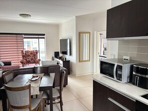 Ein modernes Wohnzimmer mit Sofa, Esstisch und Stühlen, einem Flachbildfernseher, einer Küche mit Mikrowelle und Kaffeemaschine sowie einem großen Fenster mit Jalousien.