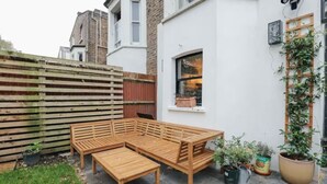 Een houten bankje en tafel staan in een tuin met een witte muur, een raam en planten in potten.