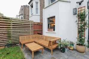 Ein Holzbänkchen und Tisch in einem Gartenbereich mit weißer Wand, Fenster und Topfpflanzen.