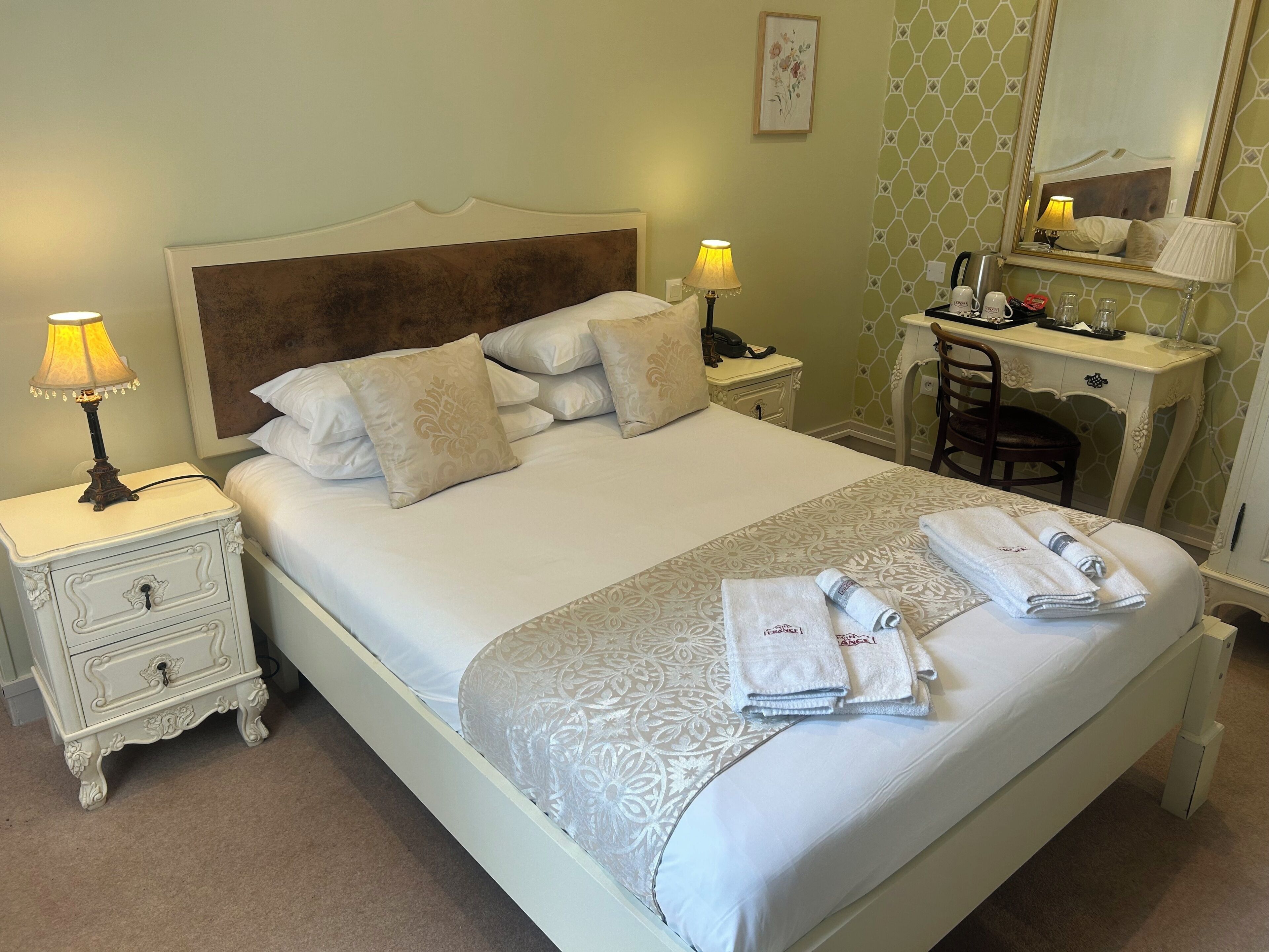 A neatly made bed with a patterned bedspread, two bedside lamps, a nightstand with drawers, a desk with a chair, and a mirror on the wall.