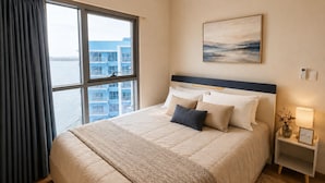 Habitación de hotel con una cama grande, vista a la ciudad y una mesita de noche con lámpara y cartel de bienvenida.