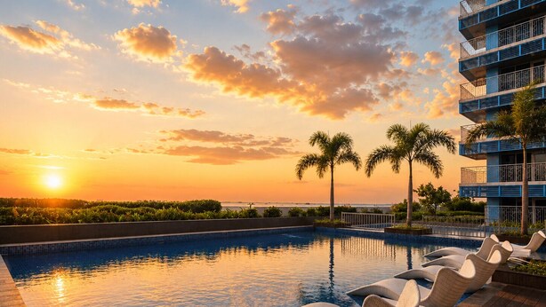 Una piscina con sillones de descanso, vista al atardecer y un edificio con balcones.